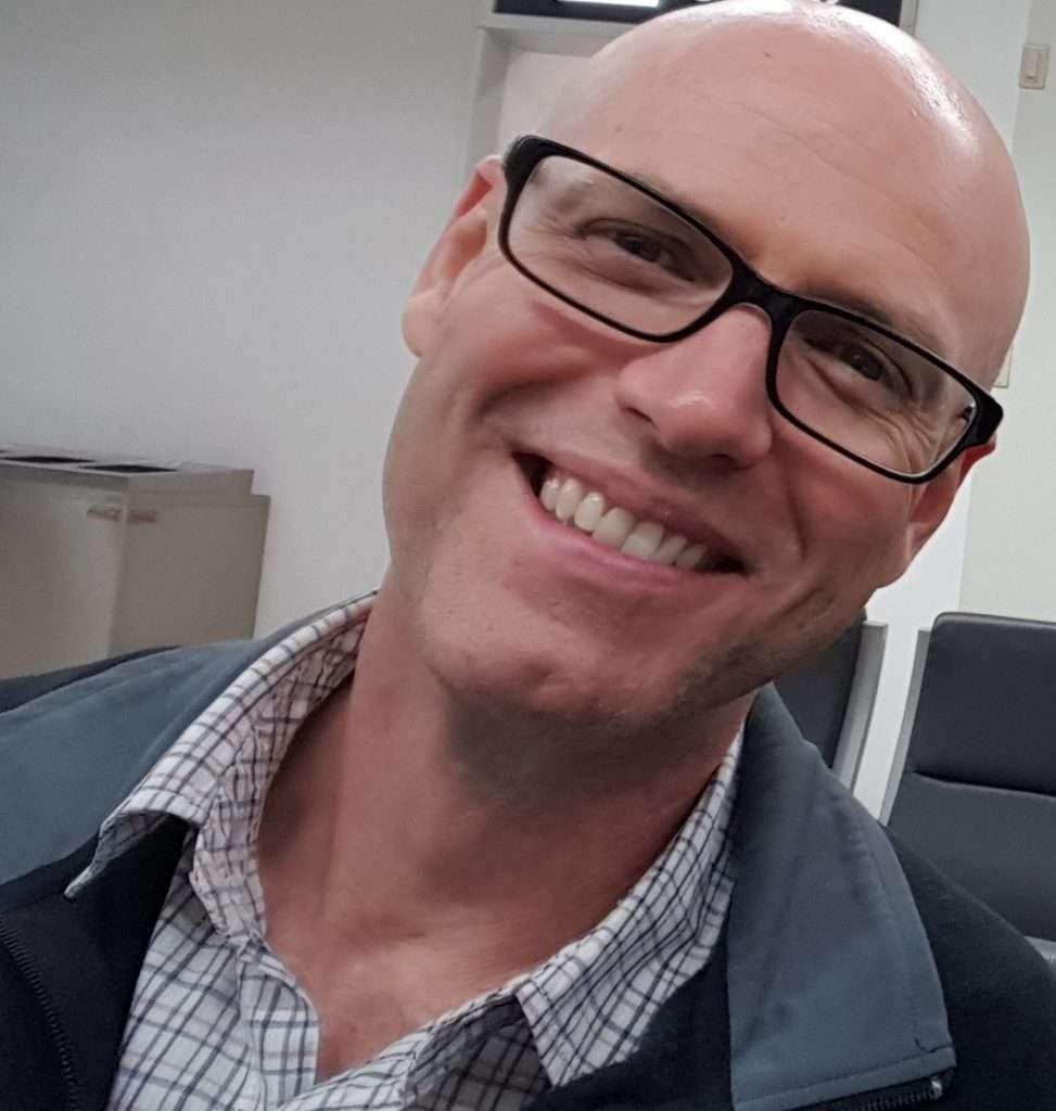 Glenn Weller portrait photo, smiling, wearing rectangular, black-framed glasses and a collared shirt beneath a black fleece jumper