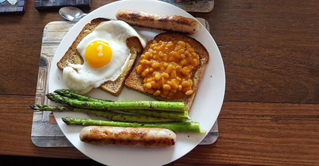 A breakfast fry up on a white plate: Fried egg on toast, baked beans on toast, two sausages, four asparagus stalks