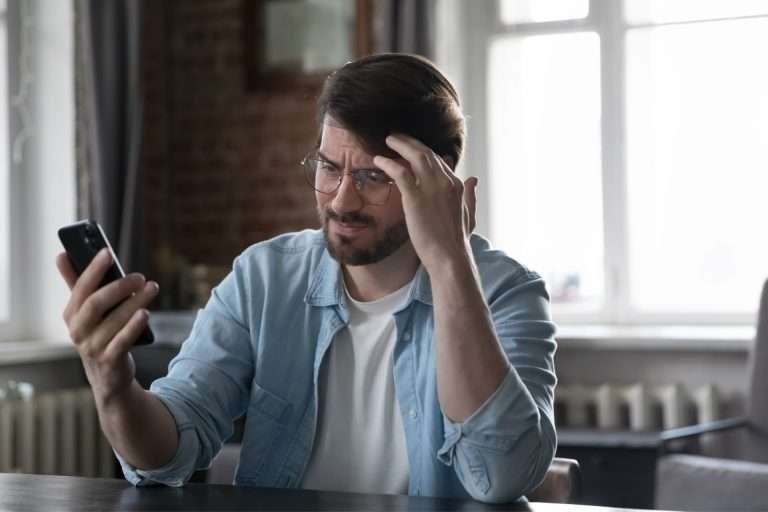 Concerned man with beard and glasses stares at a mobile phone with his elbows propped on a desk