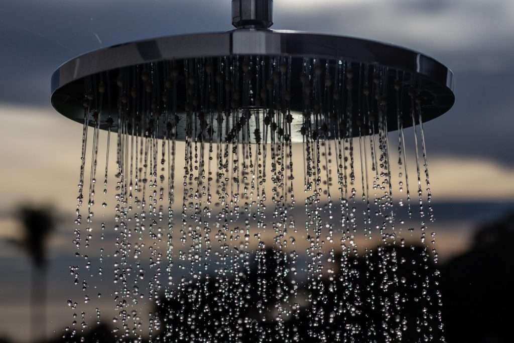 Close up of large silver rain shower head with water drops falling from it against a blurred background