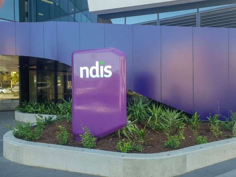 A grape-coloured sign sits in a garden bed before an office building with the letters ndis written on it in white