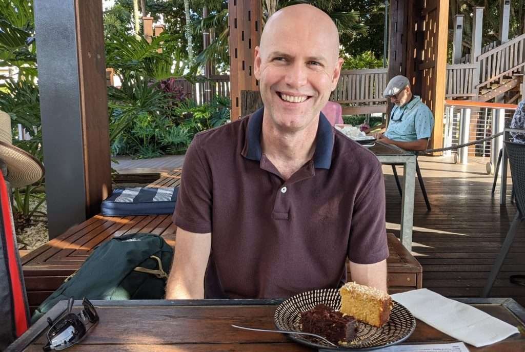 Glenn witting at a wooden table in a café with cake on a plate in front of him.