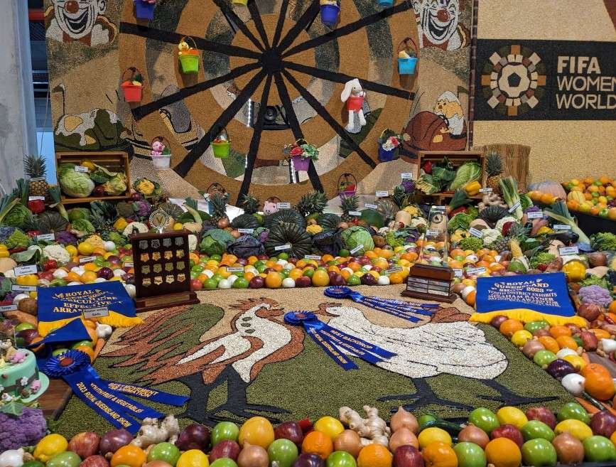 A wide assortment of fruit and vegetables interspersed with carpet-style images of chickens and clowns with a large dart board on the wall at the back is designed in a way to celebrate the Ekka.