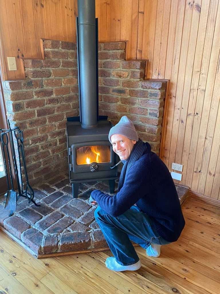 Glenn crouching before the free-standing fireplace admiring the fire he has just made.