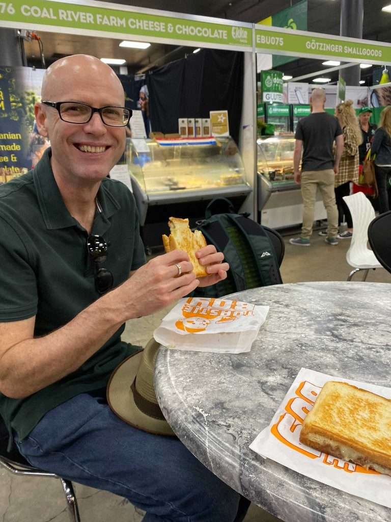 Glenn sitting at a table in the Woolworths Pavilion eating a toasted cheese sandwich. And toasted cheese sandwich rests on the table.
