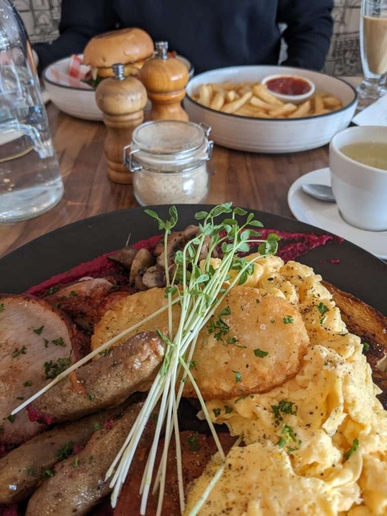 A cluttered café table with a full plate breakfast, tea cup, sugar bowl, bottle of water, pepper and salt shaker, a bowl of hot chips, and a hamburger on it.