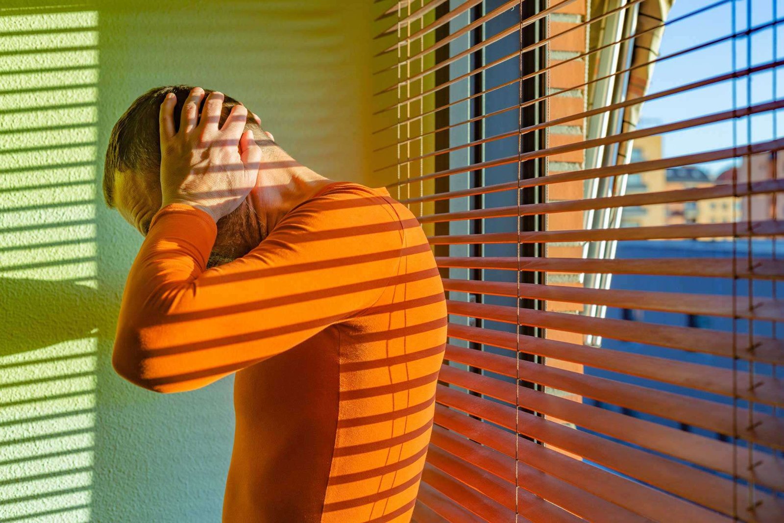 Distressed man with hands on head wearing an orange long-sleeved shirt standing by a window with venetian blinds