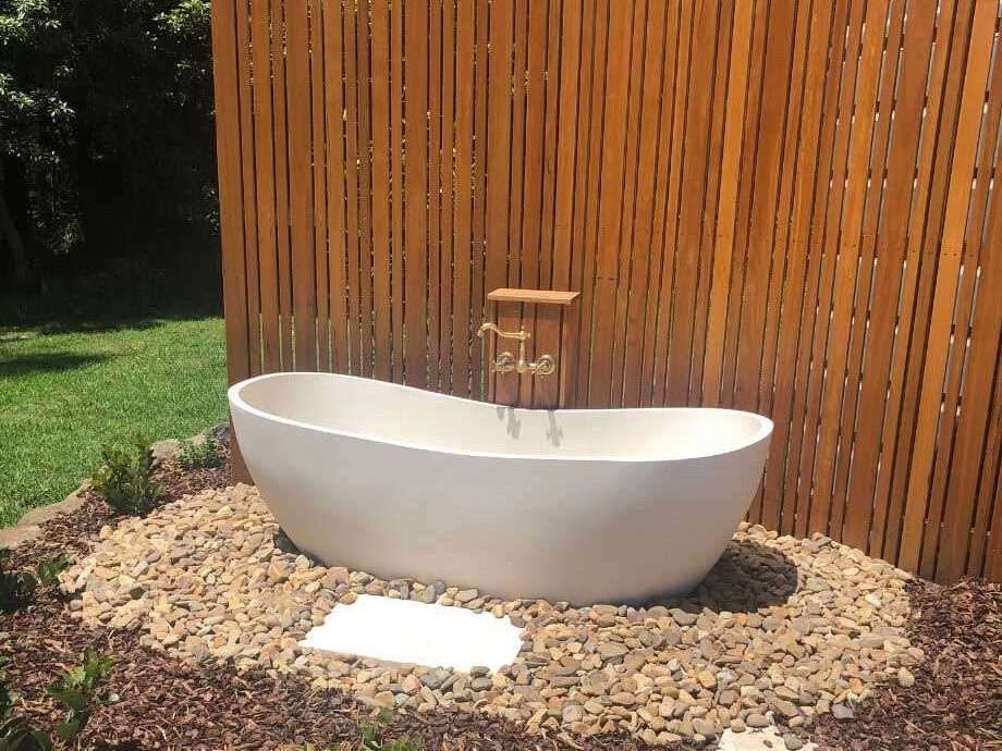 A white bathtub resting on stones outside in a garden behind an oil-stained wooden screen