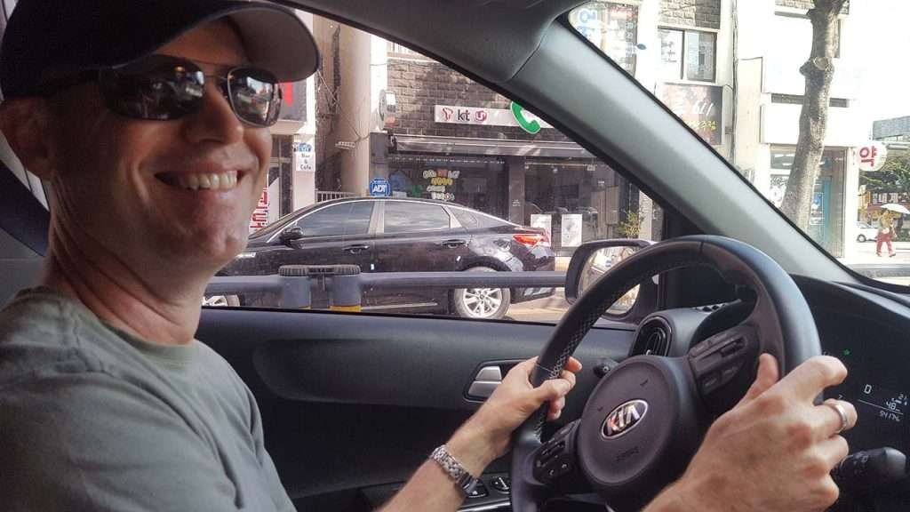 Glenn sitting behind the wheel of a car, smiling for the camera, with a South Korean streetscape in the background